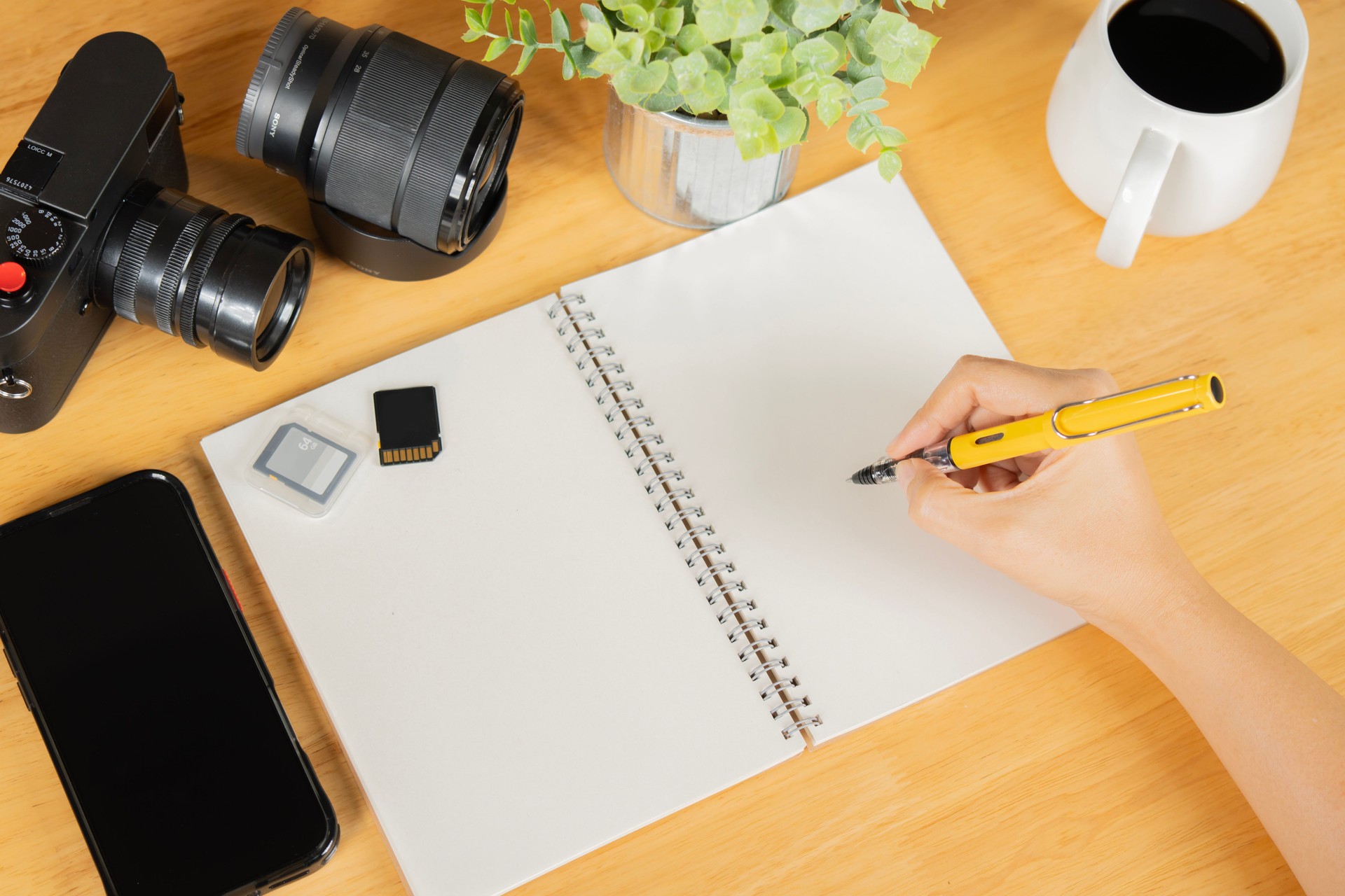 A person writing in a notebook with camera, memory cards, phone and lens on wooden table.
