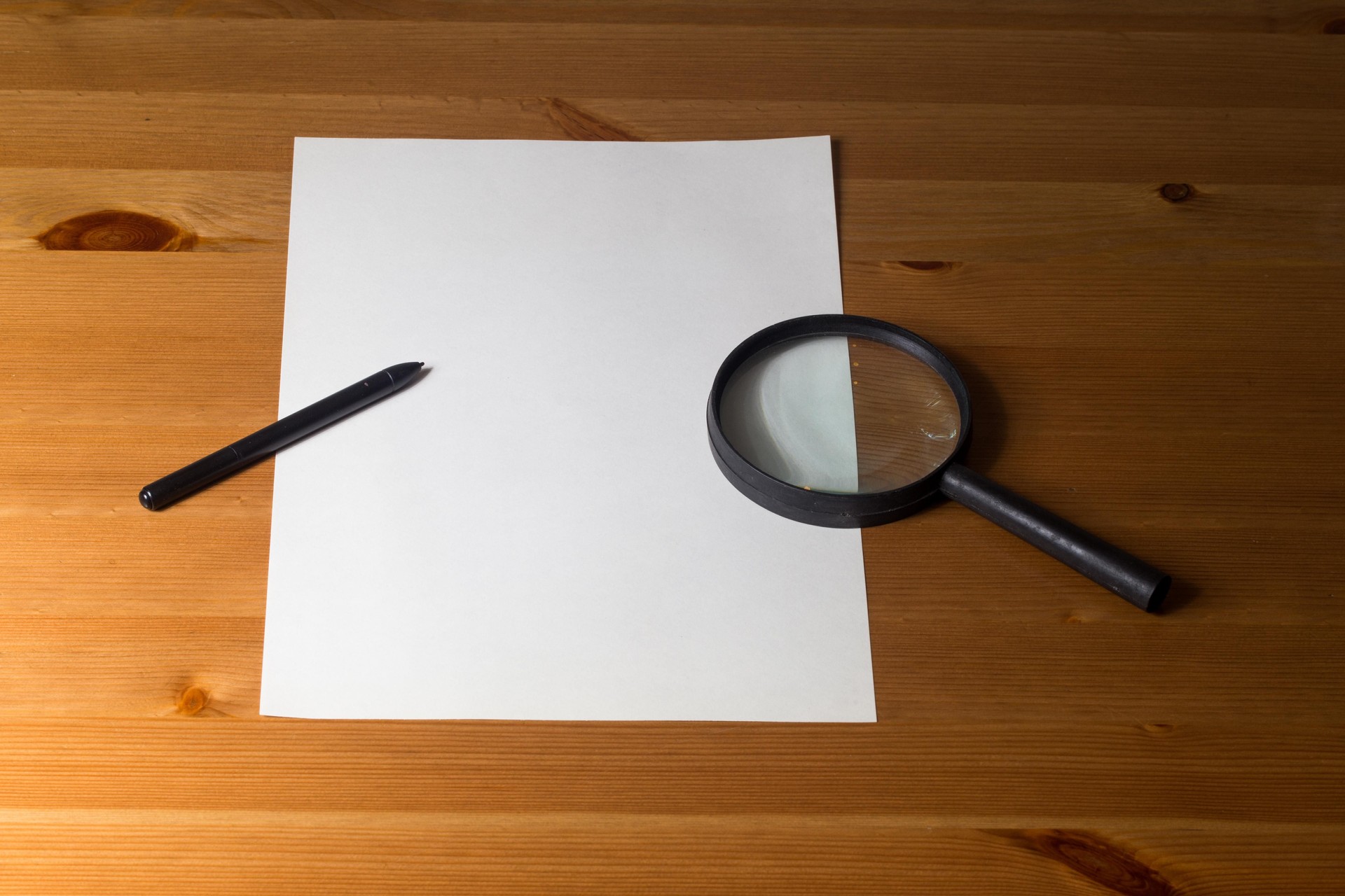 A magnifying glass, a pen, and a blank sheet of paper on a wooden table